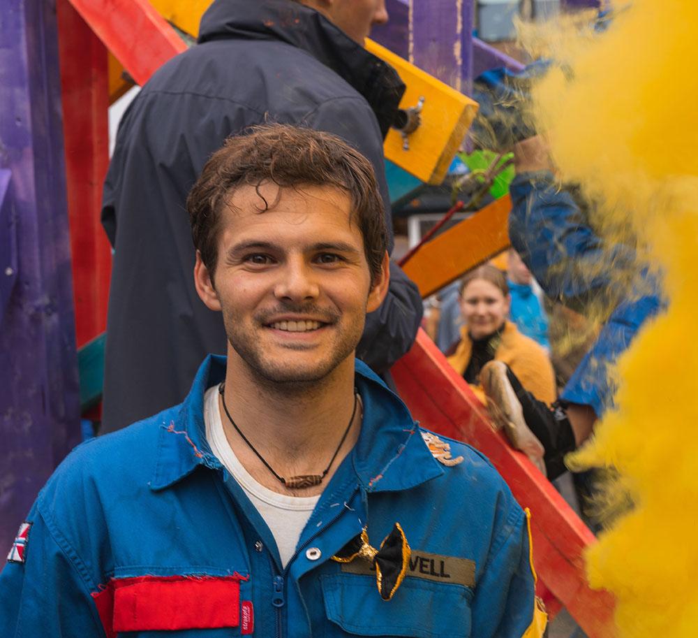 En mann med brunt hår og stort smil står i blå kjeldress foran en fargerik bakrunn. Foto