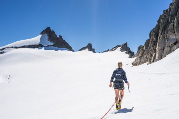 Jente med NTNU-jakke (bakfra) går på snøkledd fjell. Foto. 