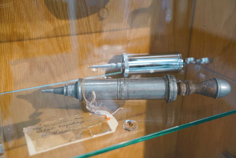 To klystersprøyter liggende på glasshylle i en utstillingsmonter.