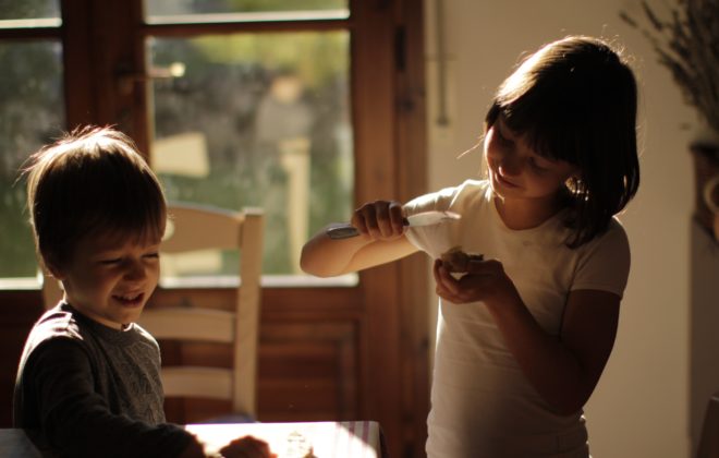 Gutt og jente som smører mat ved et kjøkkenbord i morgenlys. Foto.