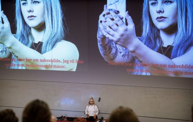 Bilde av en forelesning der en person holder foredrag, i bakgrunnen et bilde av Mia Landsem med telefon i hånda. Foto: Adresseavisen / Rune Petter Ness