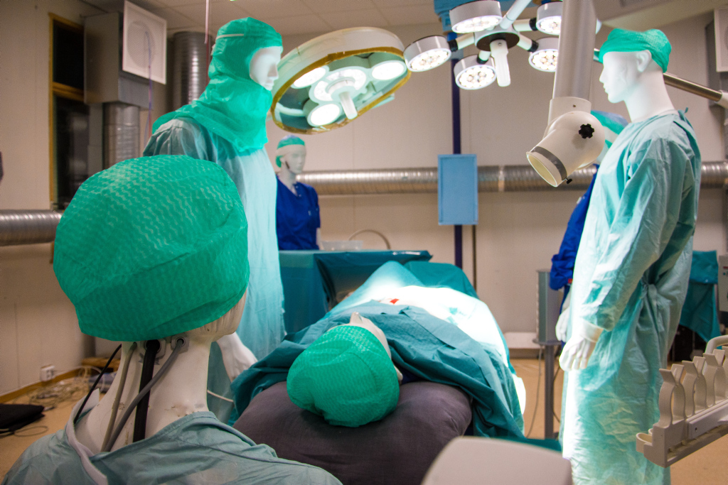 dukker med legefrakker og utstyr rundt et operasjonsbord. foto