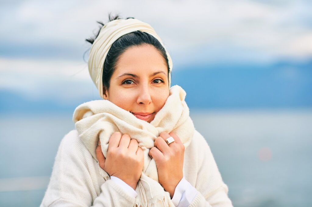 Outdoor portrait of young latin woman posing by lake or sea, warmself with big scarf