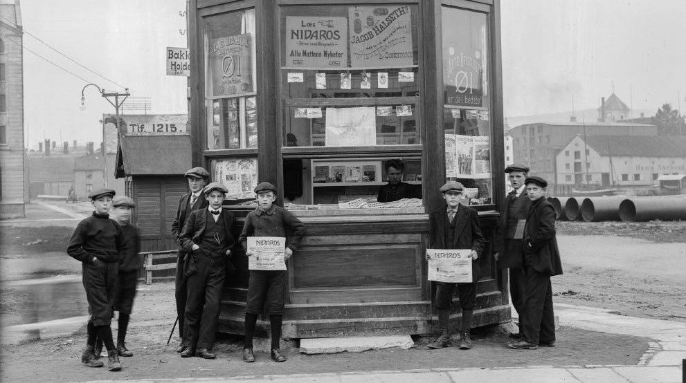 Newspaper stand with boys standing outside holding newspapers