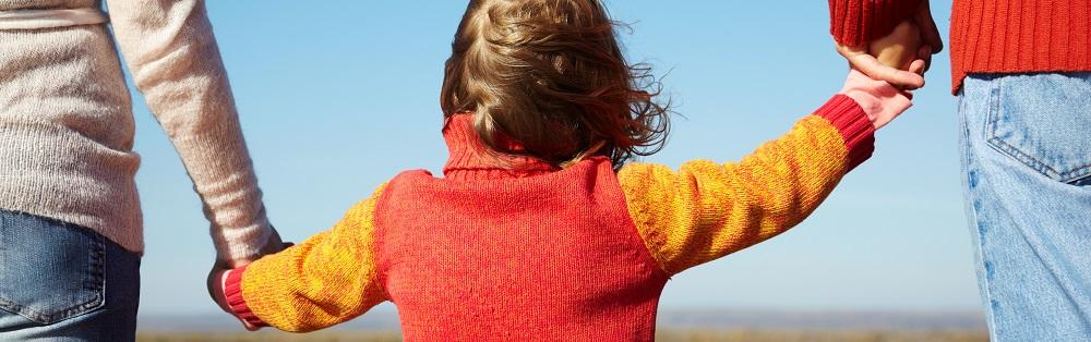 Et barn i strikket trøye holdere hender mellom to voksne som går. Sees bakfra