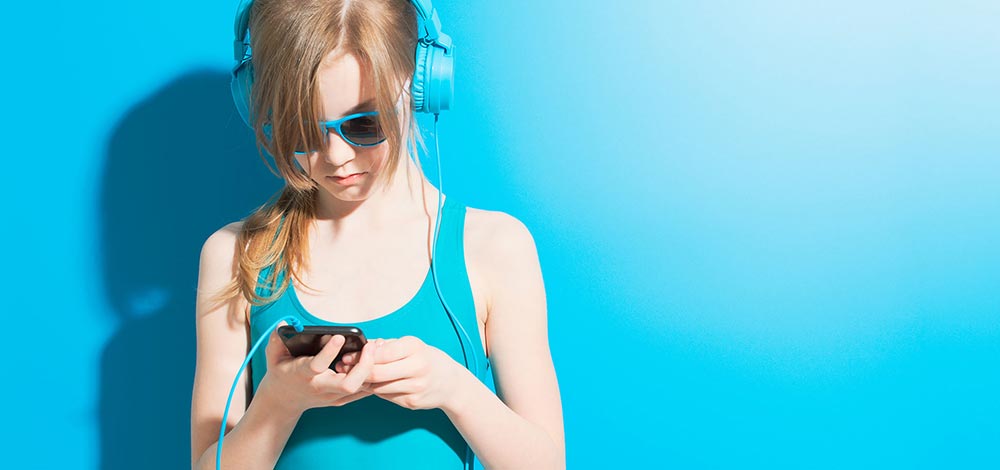 Jente med hodetelefoner som ser på mobiltelefon på blå bakgrunn, foto
