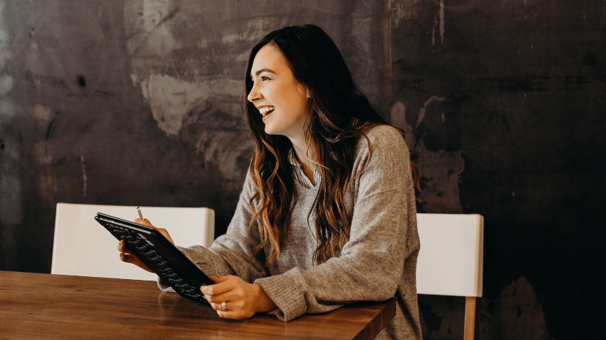 En smilende kvinne sitter med en flipbar PC i hendene ved et trebord og ser bort på noen andre. Bak henne er en grå vegg. Foto