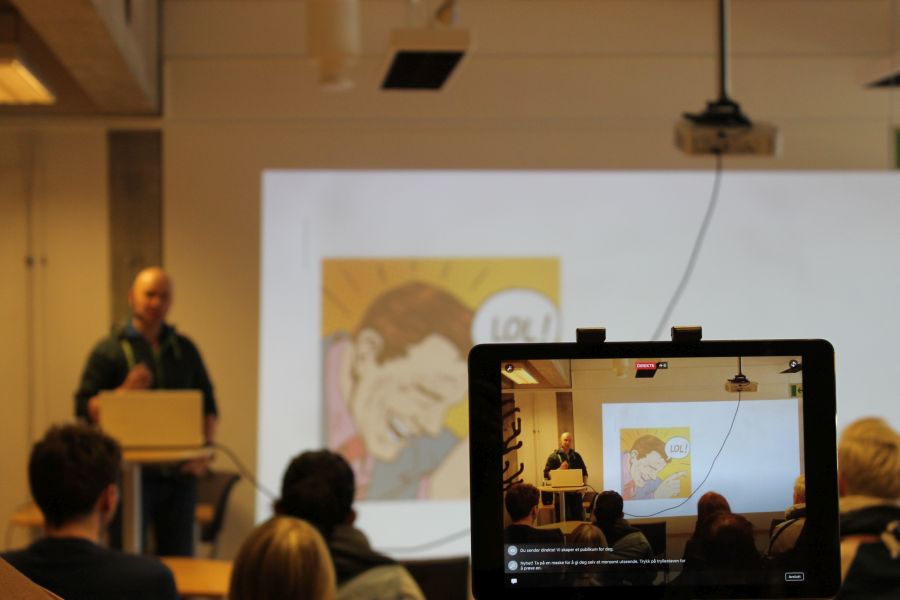 En presentasjon i et klasserom blir tatt opp med mobil. Foreleser ved podiet, publikum i stoler, lerret med tegneseriebilde
