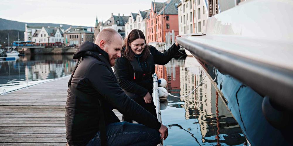 To studenter sitter på huk på brygge og sjekker ut lystbåt i Ålesund sentrum 