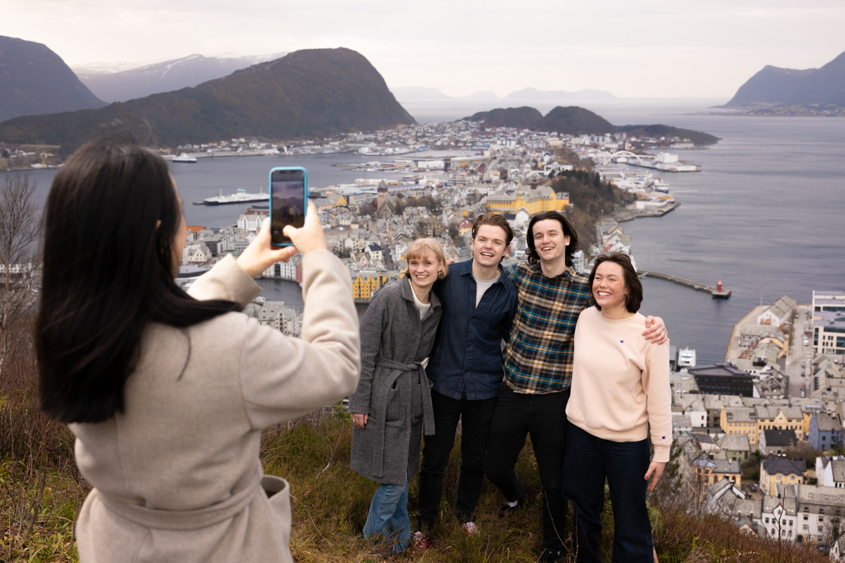 En student står og tar bilde av fire smilende studenter med øygården i Ålesund bak seg. Foto. 