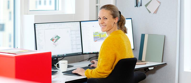 Smilende kvinne sittende ved kontorarbeidsplass med to skjermer. Foto.