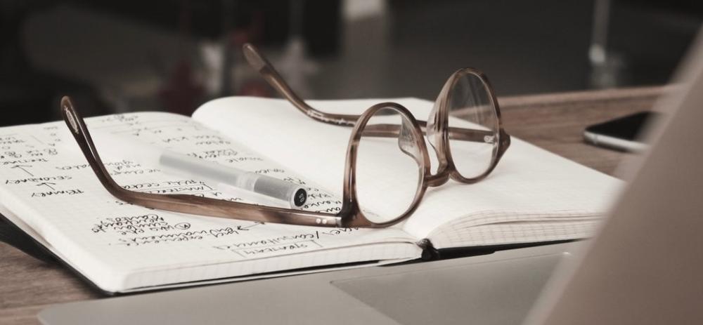 Et par briller ligger på en oppslått bok foran en bærbar pc. Bilde