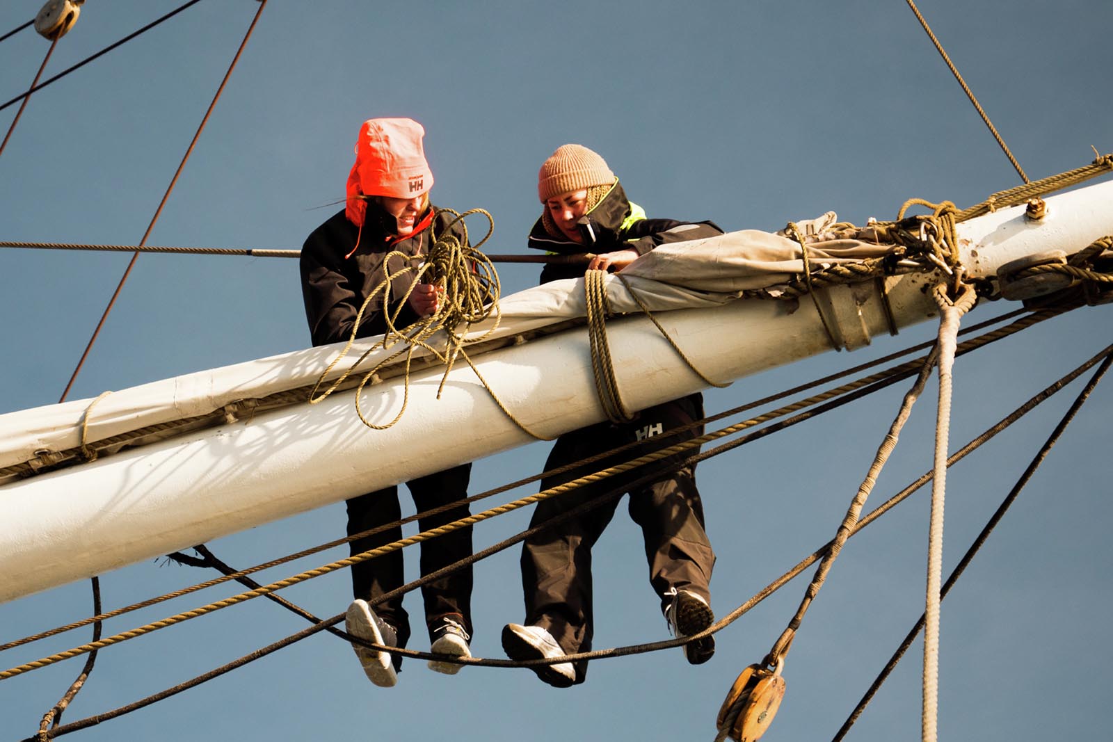 To personer står på tau og jobber med andre tau som er surret rundt en mast