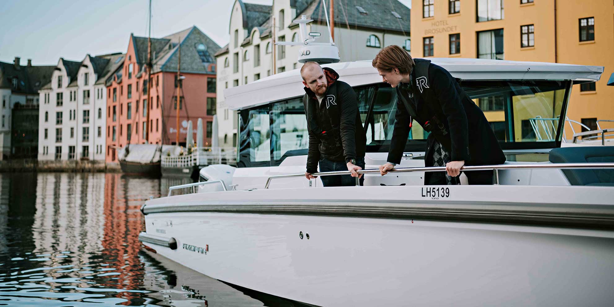 To studenter lener seg over ripa på en lystbåt på havna i Ålesund