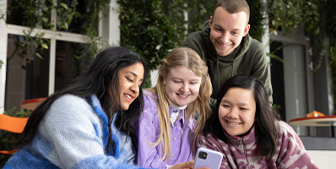 En smilende student viser noe på mobilen for tre andre smilende studenter innendørs 