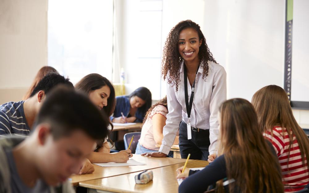 Smilende kvinnelig lærer i klasserom hvor ungdommer sitter bøyd over skolearbeid