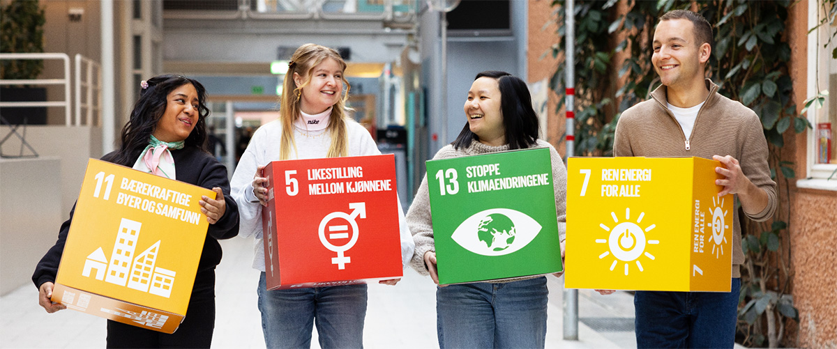 Fire smilende studenter bærer på store fargerike blokker med bærekraftsmål. Fra venstre står det bærekraftsmål 11: Bærekraftige byer og samfunn. Etterfulgt av 5 Likestilling mellom kjønnene, 13 Stopp klimaendringene og 7 Ren energi for alle. Foto.