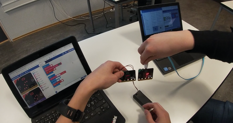 To elever som viser fram micro:bits foran PC-skjermer med programmering på. Foto.
