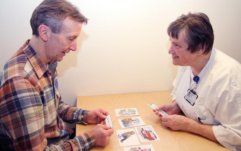Kvinnelig logoped sitter sammen med mannlig pasient. De driver språktrening ved et bord med forskjellige kort med illustrasjoner på. 
