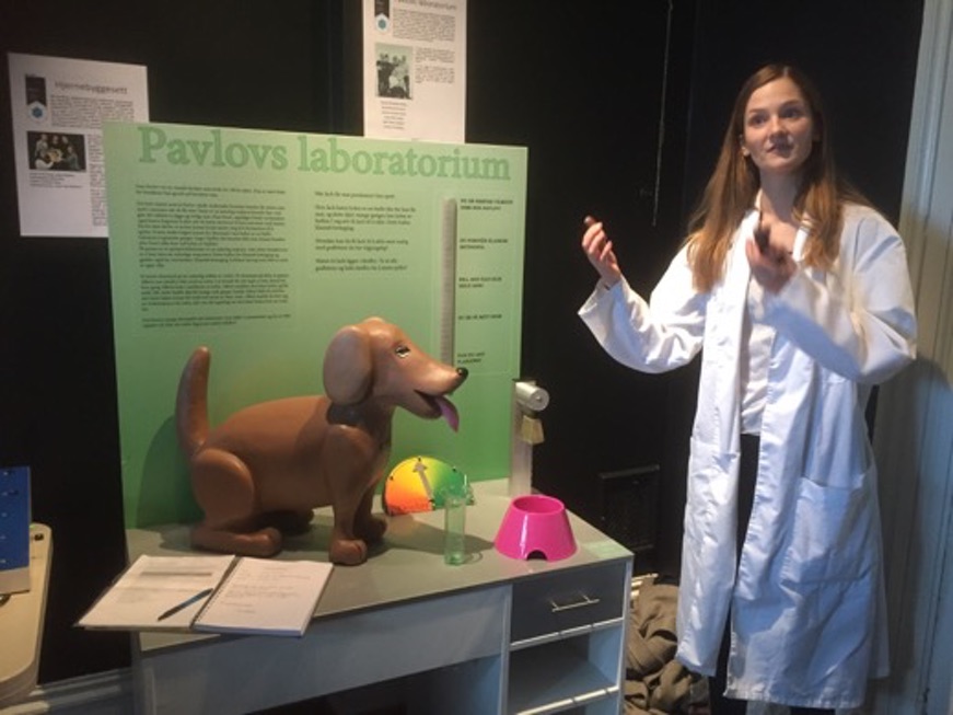 Kvinnelig student viser fram og forteller om "Pavlovs laboratorium". En robothund står på bordet.Bilde.