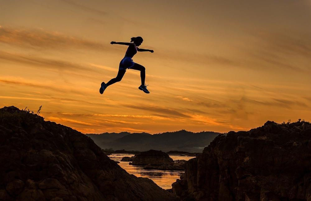 Kvinne hopper mellom fjelltopper i solnedgang.
