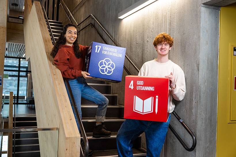 2 glade studenter, med bokser av bærekraftmålene, på campus i Gjøvik. Foto