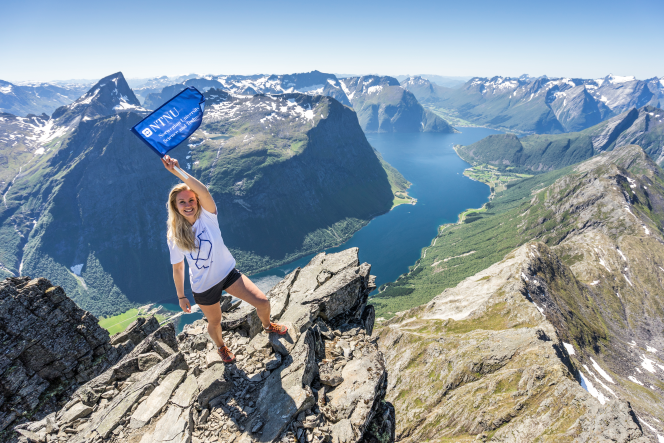 Smilende jente med NTNU-flagg på fjell. Utsikt i bakgrunn. Foto.